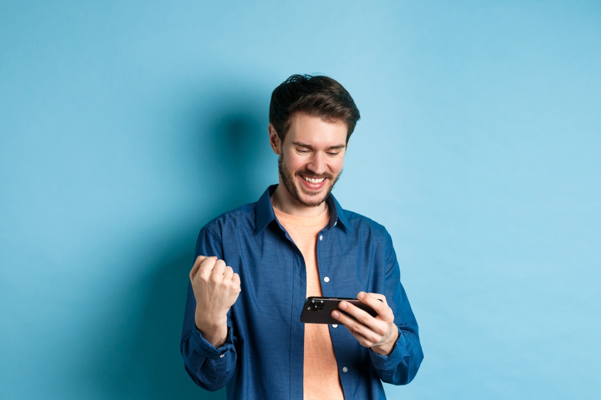 Homme sourit et serre le poing de joie, smartphone dans l'autre main, car forfait 20 Go à 6€ chez Lebara sur réseau d'Orange